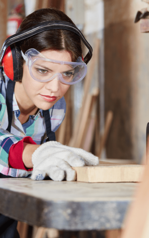 Female carpenter wearing safety equipment measures a board for cutting