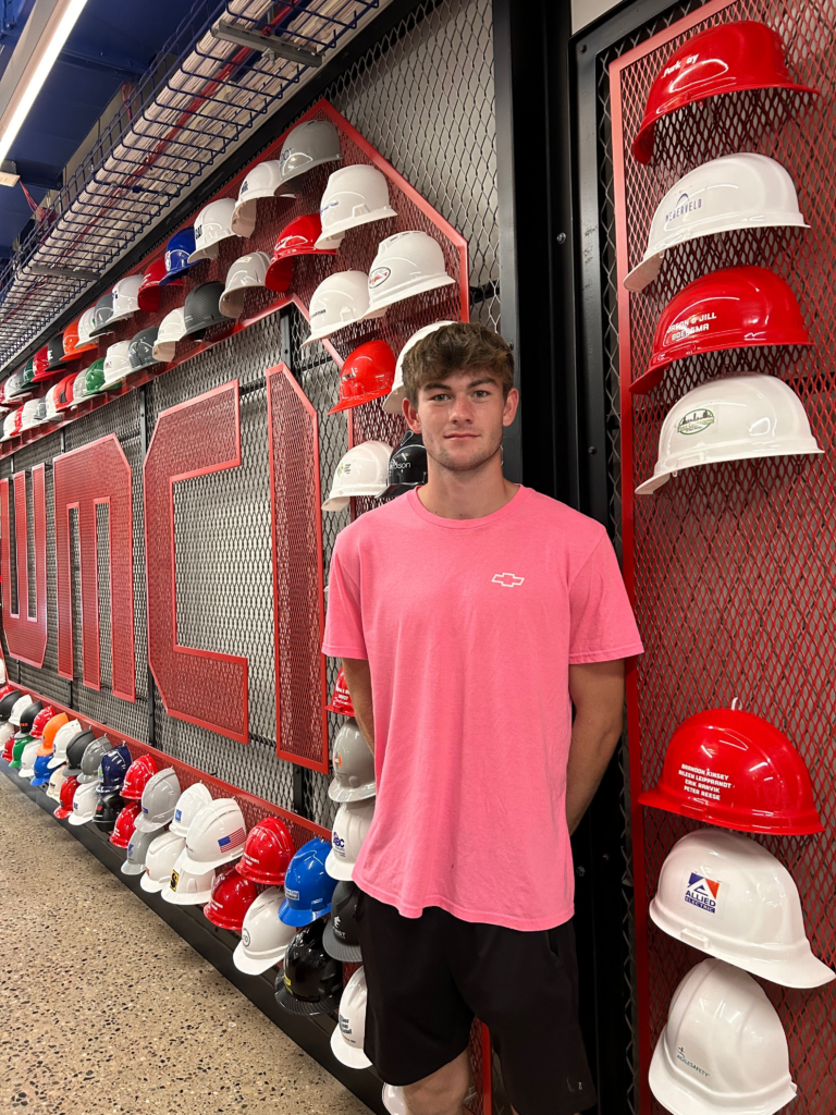 Isaac VanderMolen - 2025 recipient of the Norm Brady Scholarship, stands beside a wall of hard hats on red wire racks in a store or showroom, helmets in white, red, and other colors visible behind him.