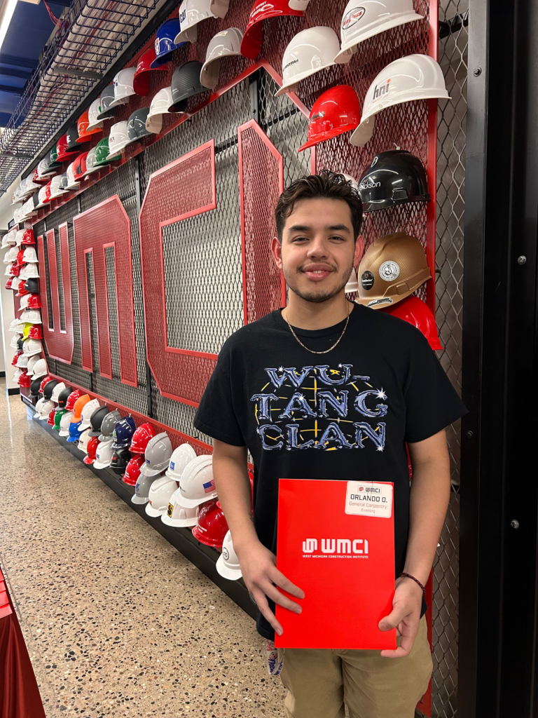 Orlando Ortiz - 2025 recipient of the WMCI general scholarship - standing in front of a wall of hard hats arranged on red mesh shelves, holding a red folder with WMCI branding.