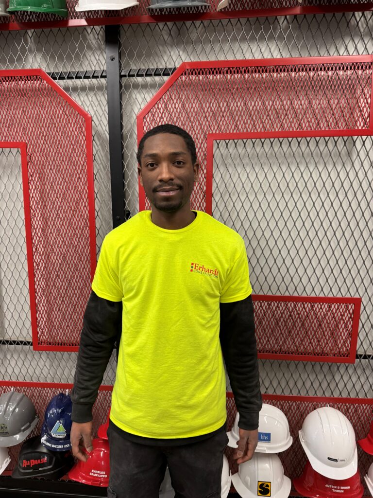Shaquille Mitchell - 2025 recipient of the WMCI general scholarship - standing in front of a wall of hard hats arranged on red mesh shelves, holding a red folder with WMCI branding.
