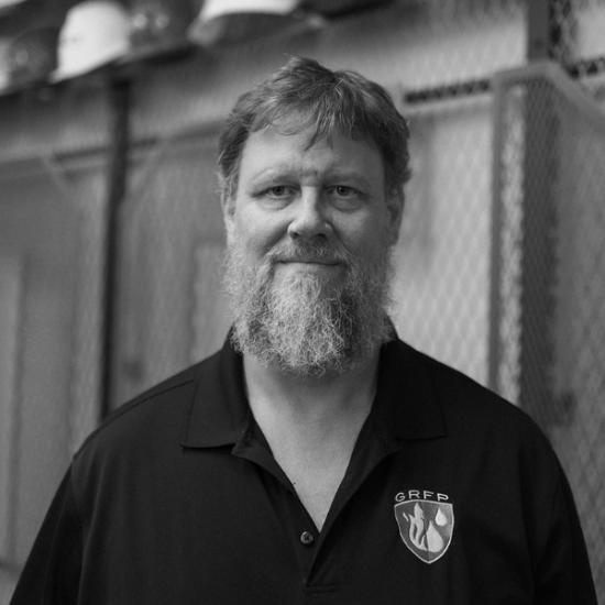 Middle-aged man with a gray beard wearing a black polo shirt with a GRFP shield logo, standing in an industrial setting with a chain-link fence in the background.